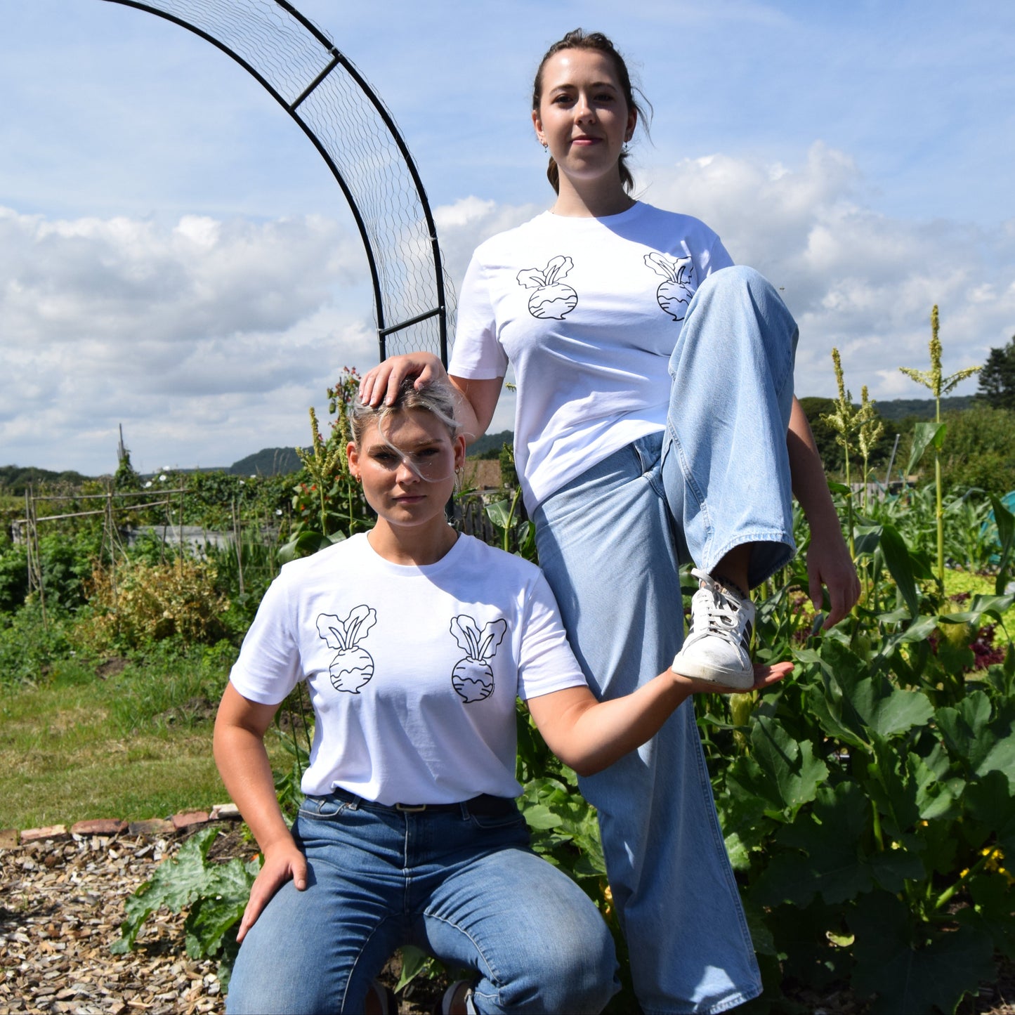 Two people in matching white t-shirts with a logo, standing in an outdoor garden setting.