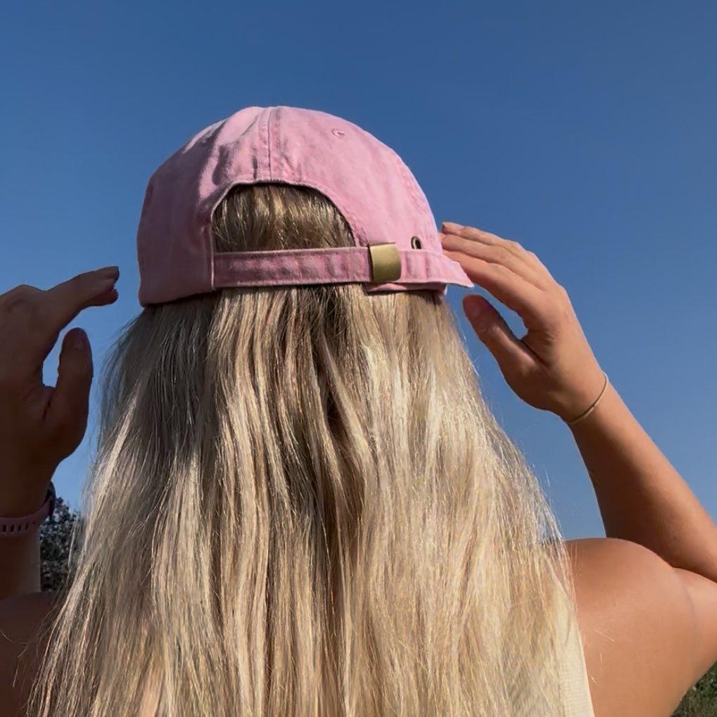 A person viewed from behind wearing a light pink baseball cap and a cream-colored tank top, standing outdoors under a clear blue sky.