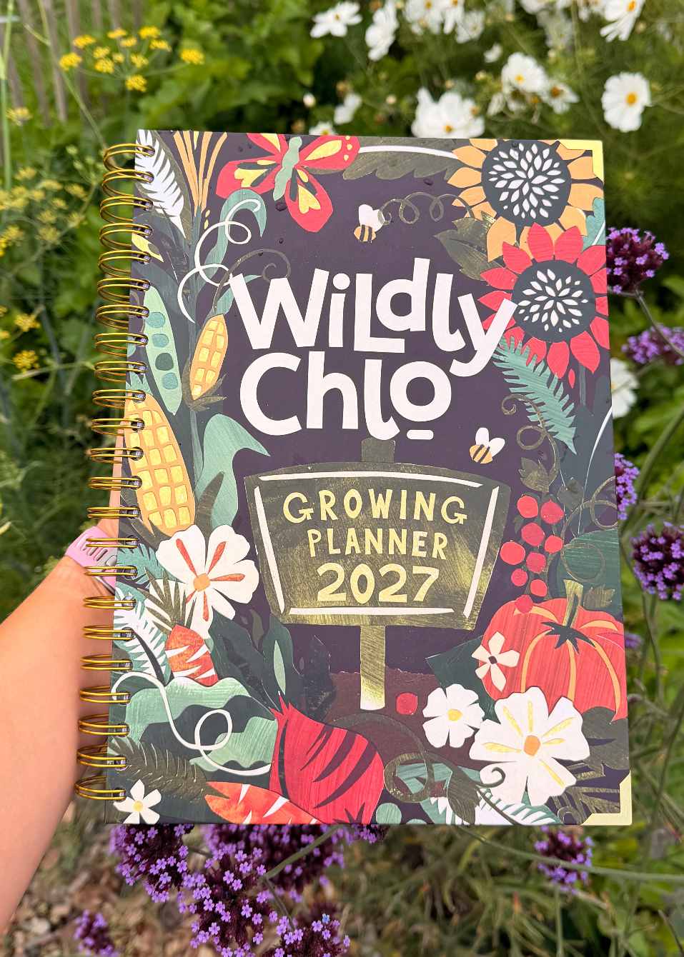 A hand holds a spiral-bound 2027 Growing Planner titled 'Wildly Chlo' against a background of green garden foliage with yellow and purple flowers. The planner cover features a colorful autumn harvest illustration with pumpkins, corn, and wildflowers.