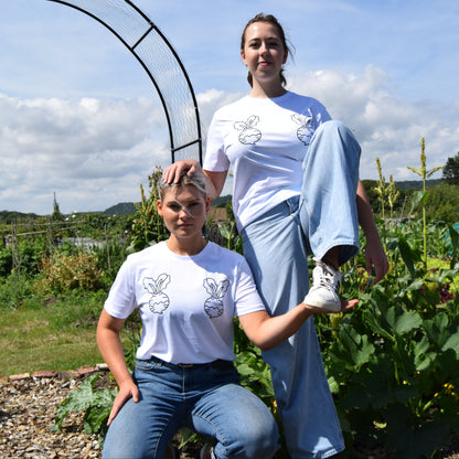 Two people in matching white t-shirts with a logo, standing in an outdoor garden setting.