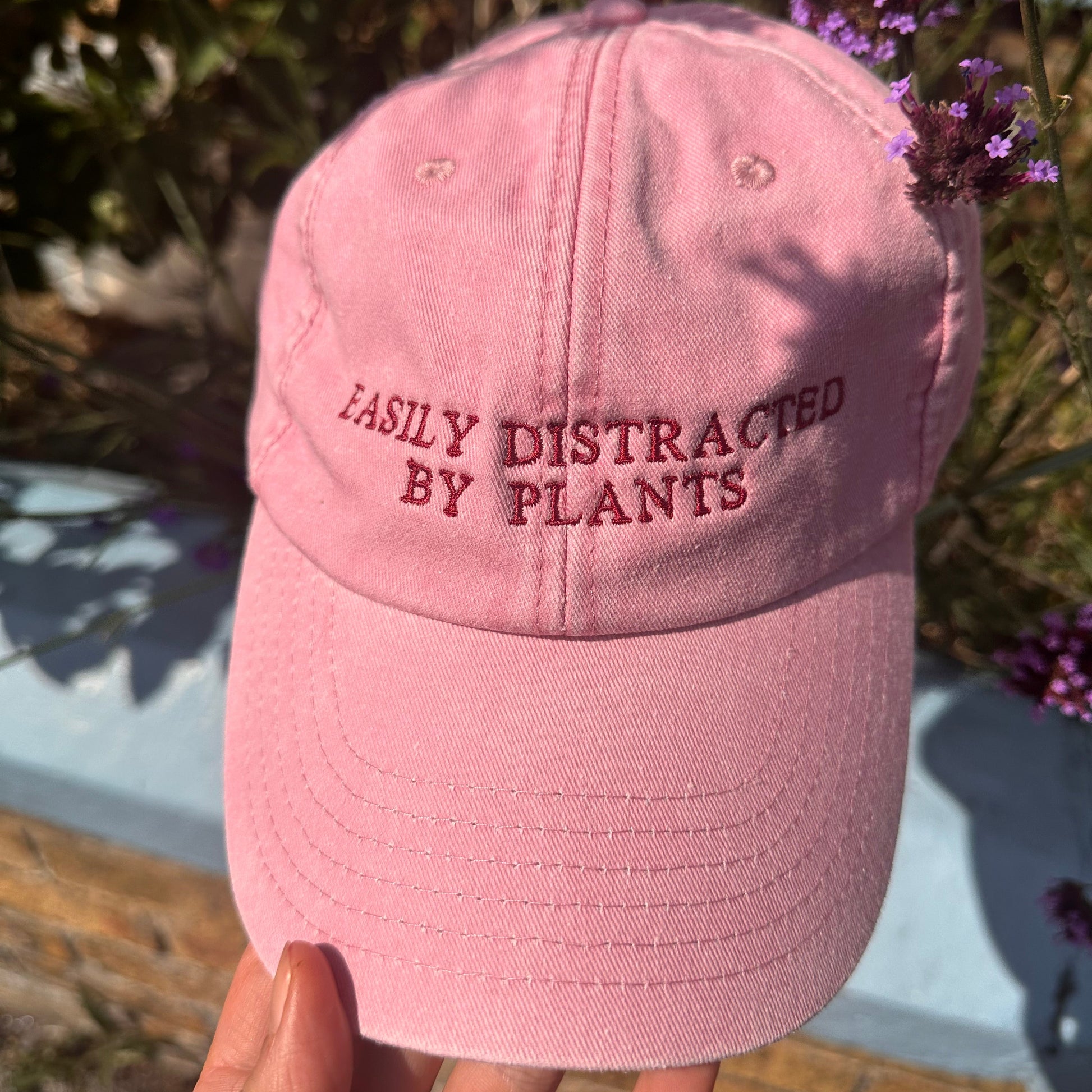 A pink baseball cap held by a hand outdoors, with the embroidered text 'EASILY DISTRACTED BY PLANTS' visible on the front panel.