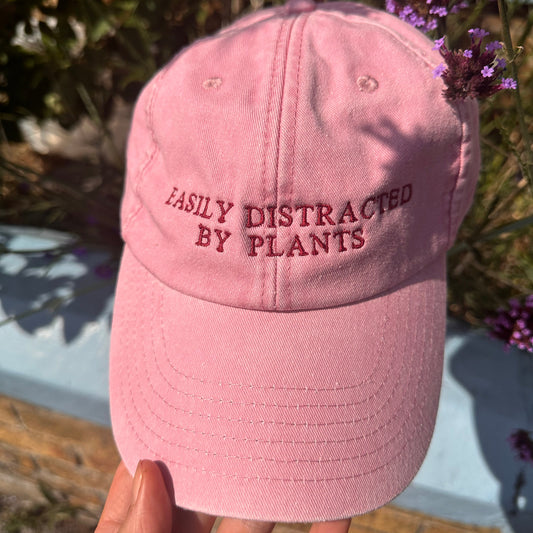 A pink baseball cap held by a hand outdoors, with the embroidered text 'EASILY DISTRACTED BY PLANTS' visible on the front panel.