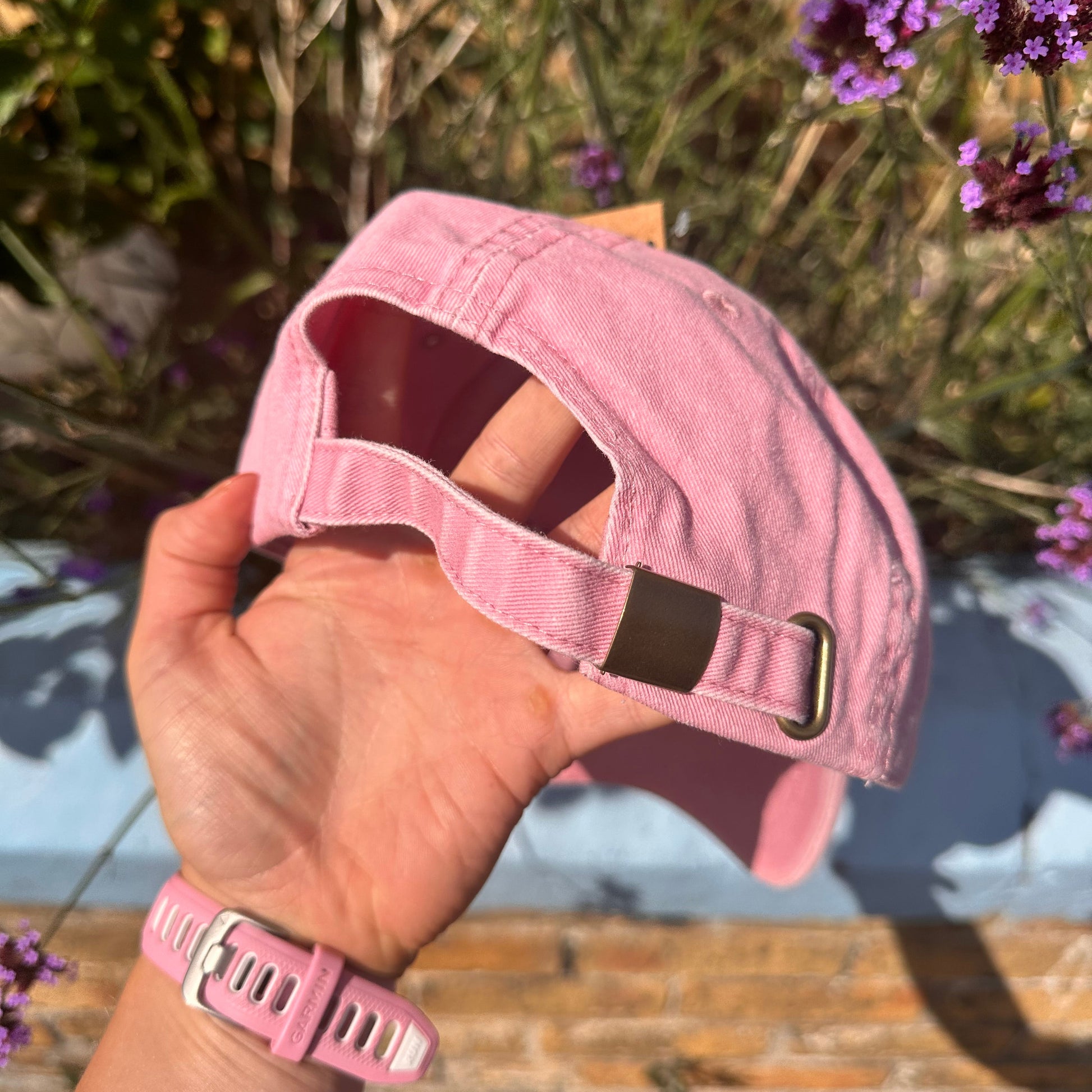 A person's hand holding a light pink baseball cap outdoors, with purple flowers and a rustic wooden surface in the background.