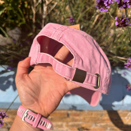 A person's hand holding a light pink baseball cap outdoors, with purple flowers and a rustic wooden surface in the background.