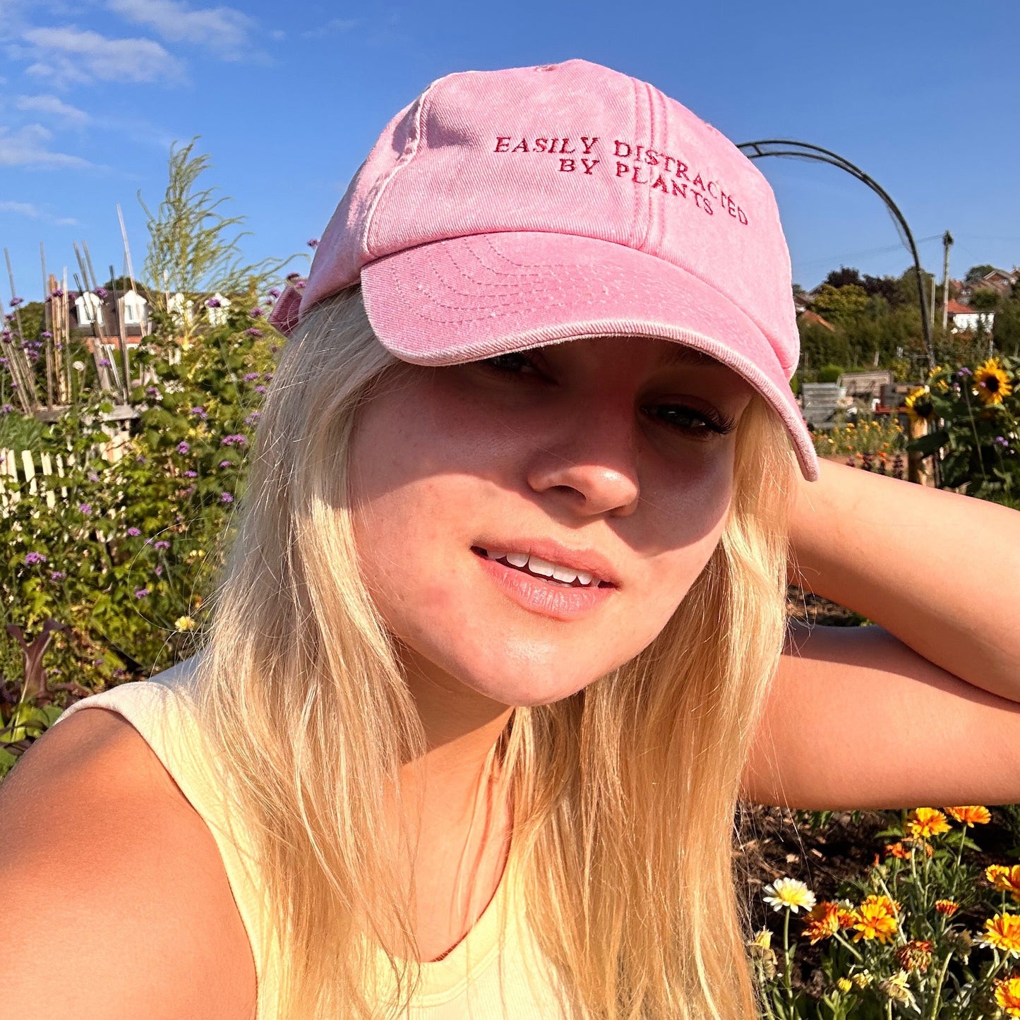 A woman with blonde hair wearing a pink baseball cap that reads 'EASILY DISTRICT BY PLANET 99' and a pale yellow ribbed tank top, posing outdoors in a sunny garden setting.