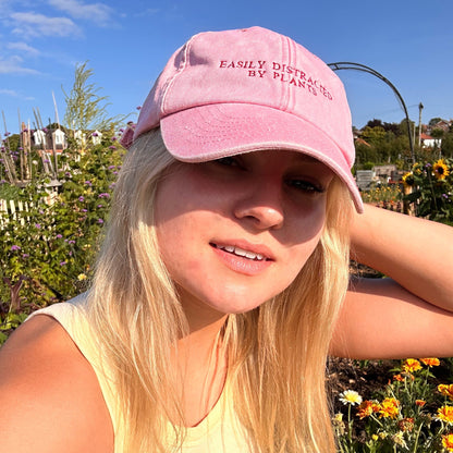 A woman with blonde hair wearing a pink baseball cap that reads 'EASILY DISTRICT BY PLANET 99' and a pale yellow ribbed tank top, posing outdoors in a sunny garden setting.