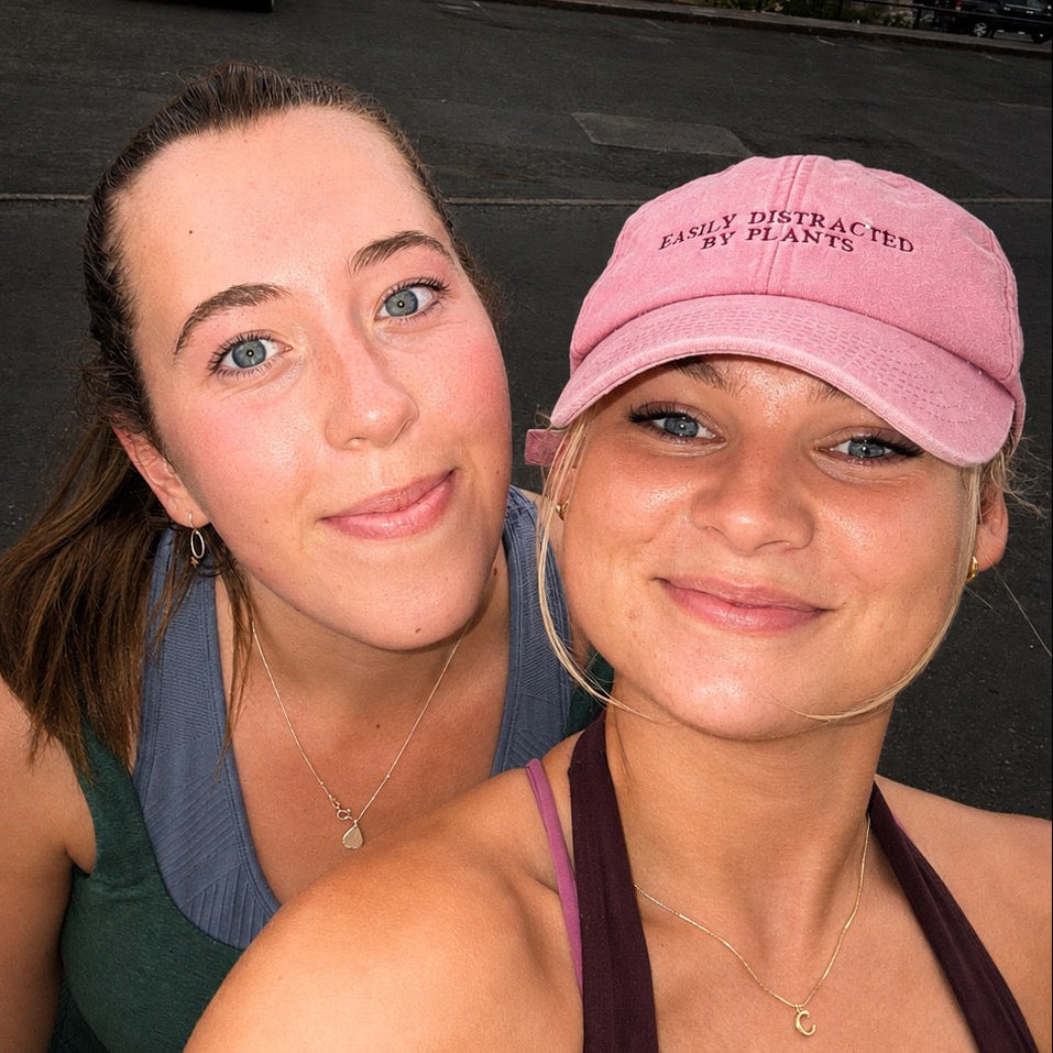 Two women taking a selfie outdoors in front of a stone building. The woman in the foreground wears a pink baseball cap with the text 'EASILY DISTRACTED BY HEARS.