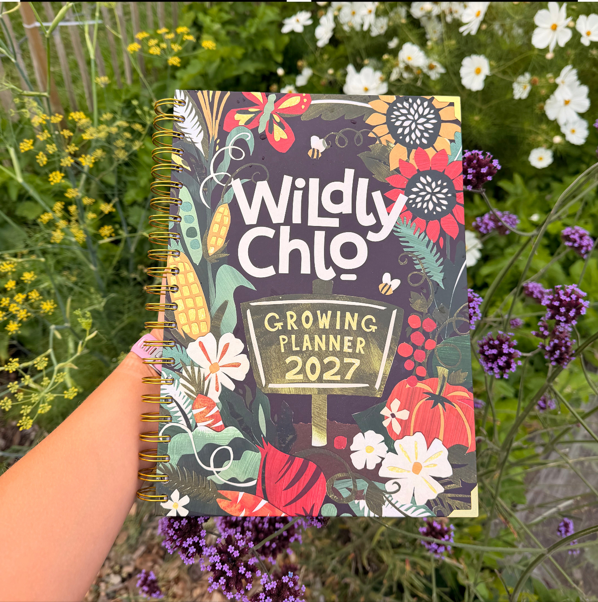 A hand holds a spiral-bound 2027 Growing Planner titled 'Wildly Chlo' against a background of green garden foliage with yellow and purple flowers. The planner cover features a colorful autumn harvest illustration with pumpkins, corn, and wildflowers.
